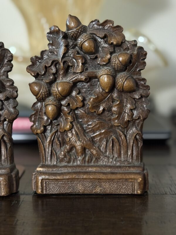 A bronze-colored metal bookend with detailed oak leaves and acorns sits on a dark wooden surface next to a Chinese Blue & White Miniature Vase, while other elegant objects appear blurred in the background.