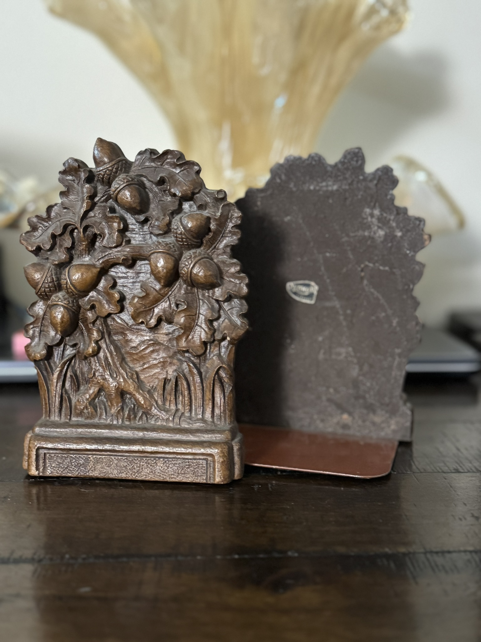 A close-up of two ornate vintage metal bookends—one with an oak leaf and acorn motif—on dark wood, with a Chinese Blue & White Miniature Vase softly blurred in the background.