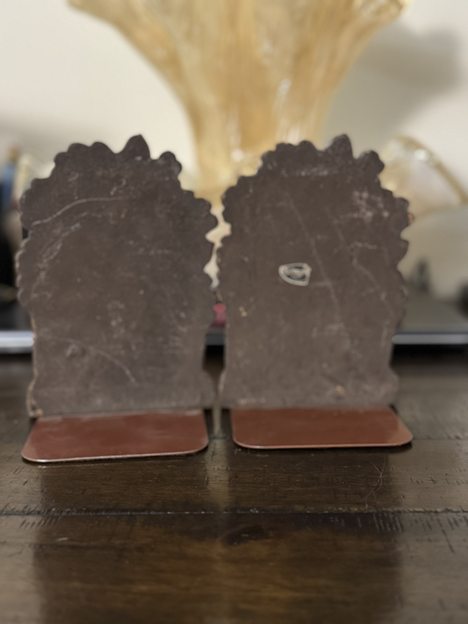 Two metal bookends with jagged edges rest on a wooden surface. The worn, dark metal has scratches and a small sticker. A Chinese Blue & White Miniature Vase is softly blurred in the background.