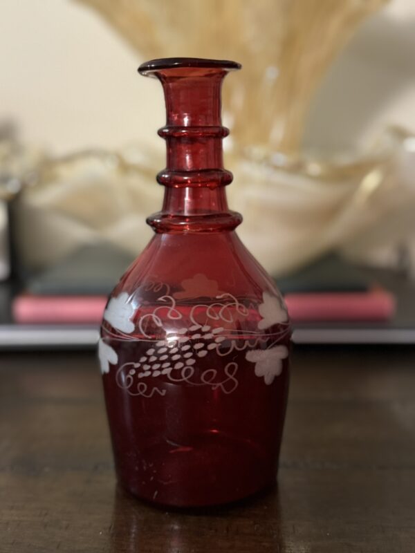 A Victorian Cranberry Glass Decanter with etched grape and leaf designs sits on a dark wooden table, its three ringed sections near the top highlighted against a softly blurred beige and gold background.