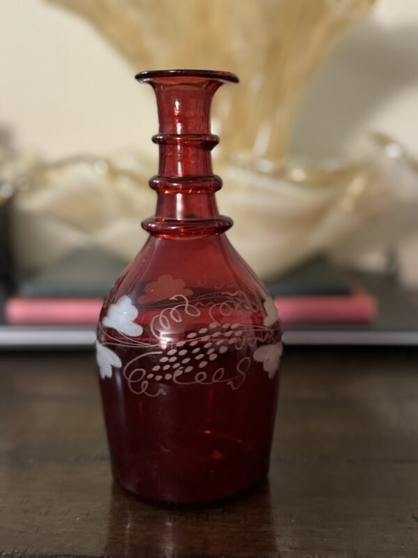 A Victorian Cranberry Glass Decanter with etched grapes rests on a dark wooden surface. Its deep red body features white grape and leaf designs, a flared lip, and three raised neck rings, with blurred golden fabric in the background.