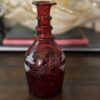 The Victorian Cranberry Glass Decanter with Etched Grapes, featuring a narrow neck and flared rim, rests on wood. Deep red glass is adorned with white etched grapes and leaves; a glass dish and two closed books blur in the background.