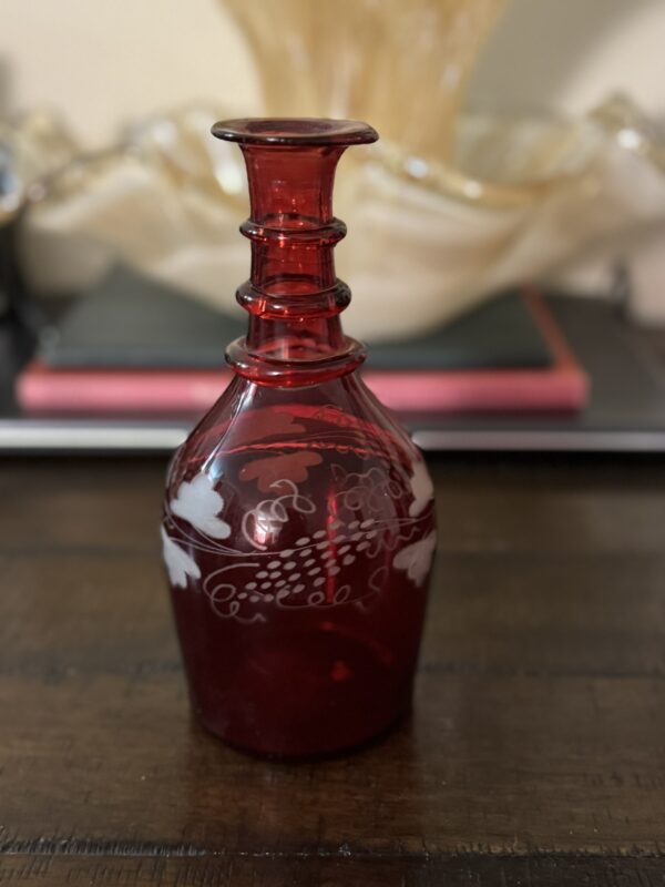The Victorian Cranberry Glass Decanter with Etched Grapes, featuring a narrow neck and flared rim, rests on wood. Deep red glass is adorned with white etched grapes and leaves; a glass dish and two closed books blur in the background.