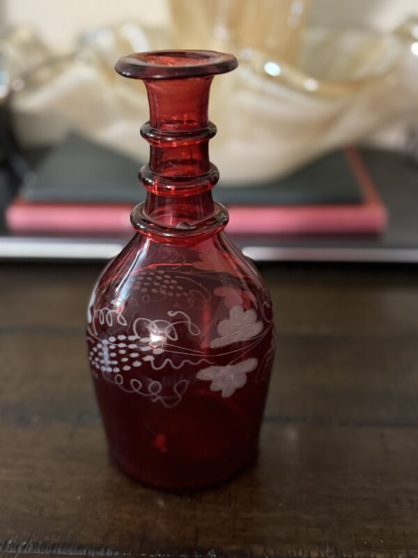 A Victorian Cranberry Glass Decanter with etched grape designs and a narrow neck sits on a wooden surface. In the blurred background, books and a wavy-edged glass bowl are visible.