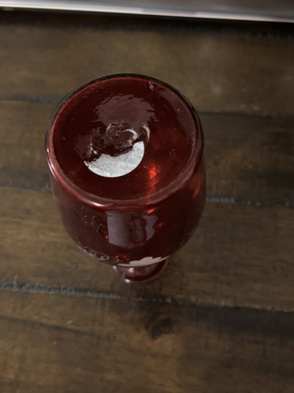 A Victorian Cranberry Glass Decanter with Etched Grapes rests upside down on a dark wooden table, displaying a partially peeled white sticker on the base and revealing clear glass beneath, with visible wood grain and table seams.