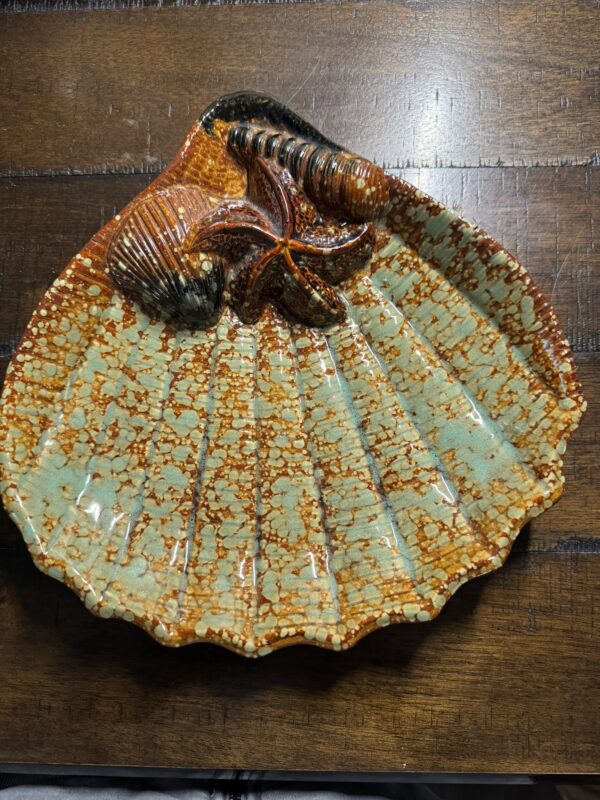 The Seashell Serving Tray is a scallop shell-shaped ceramic dish with textured ridges, glazed in orange, brown, and aqua green, and features raised clam and mussel designs near the hinge, displayed on a dark wooden surface.