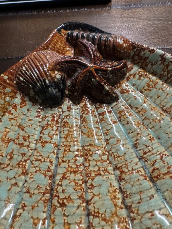 A close-up of the Seashell Serving Tray shaped like a scallop shell, with raised brown starfish and seashell designs. The ceramic tray features an earthy brown glaze with turquoise speckles and sits on a wooden surface.