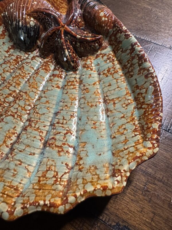 A close-up of the Seashell Serving Tray, shaped and textured like a scallop shell, with a starfish accent. The tray displays an orange, brown, and turquoise speckled glaze and rests on a wood surface with visible grain and seams.