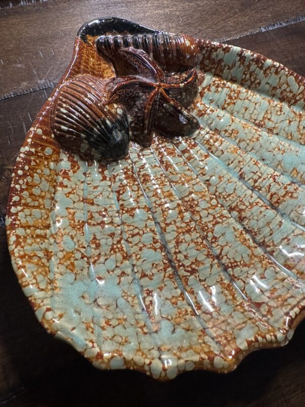 The Seashell Serving Tray, shaped like a scallop shell with speckled turquoise and rusty orange glaze, features three raised seashells—a starfish and two clamshells—at its wide end. Ideal for adding a unique touch to your table.