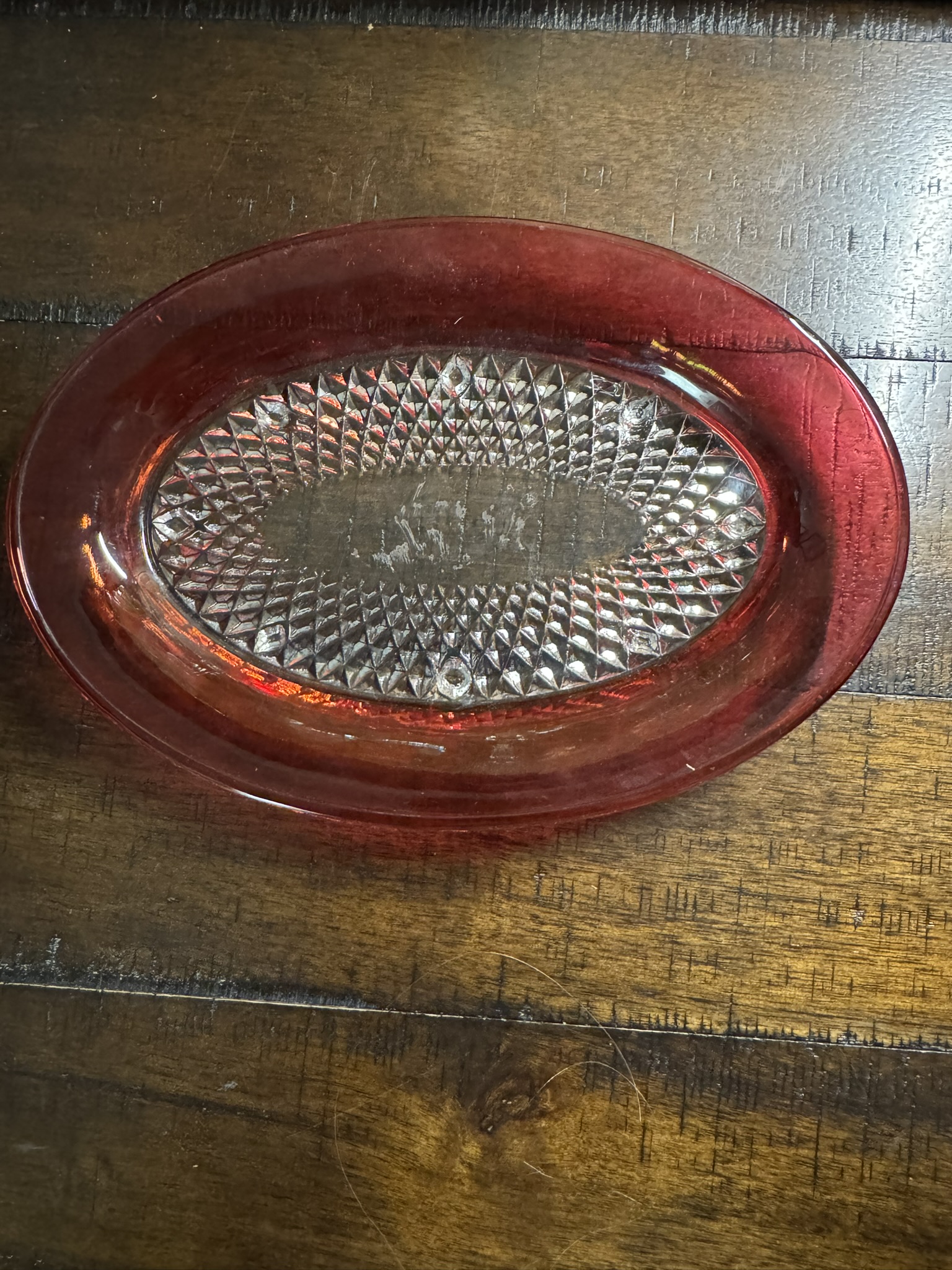 The Ruby Flash Tray, a small oval dish with a clear diamond-patterned center and a vivid red rim, rests on dark wood. Light reflects off its intricate details, contrasting the textured design with the smooth, translucent edge.