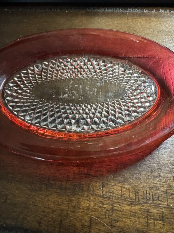 The Ruby Flash Tray, featuring a red glass dish with an oval clear textured center and diamond-cut pattern, rests on a dark wooden surface. Its translucent red edges shimmer as light highlights the intricate central design.