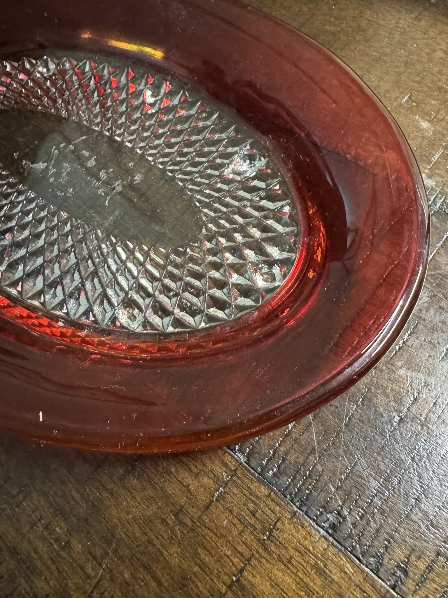 A close-up of the Ruby Flash Tray, an oval red glass dish with a diamond-textured center, sits on a brown wooden table. Light reflects off the dish, revealing some dust and showing the wood grain and seams below.