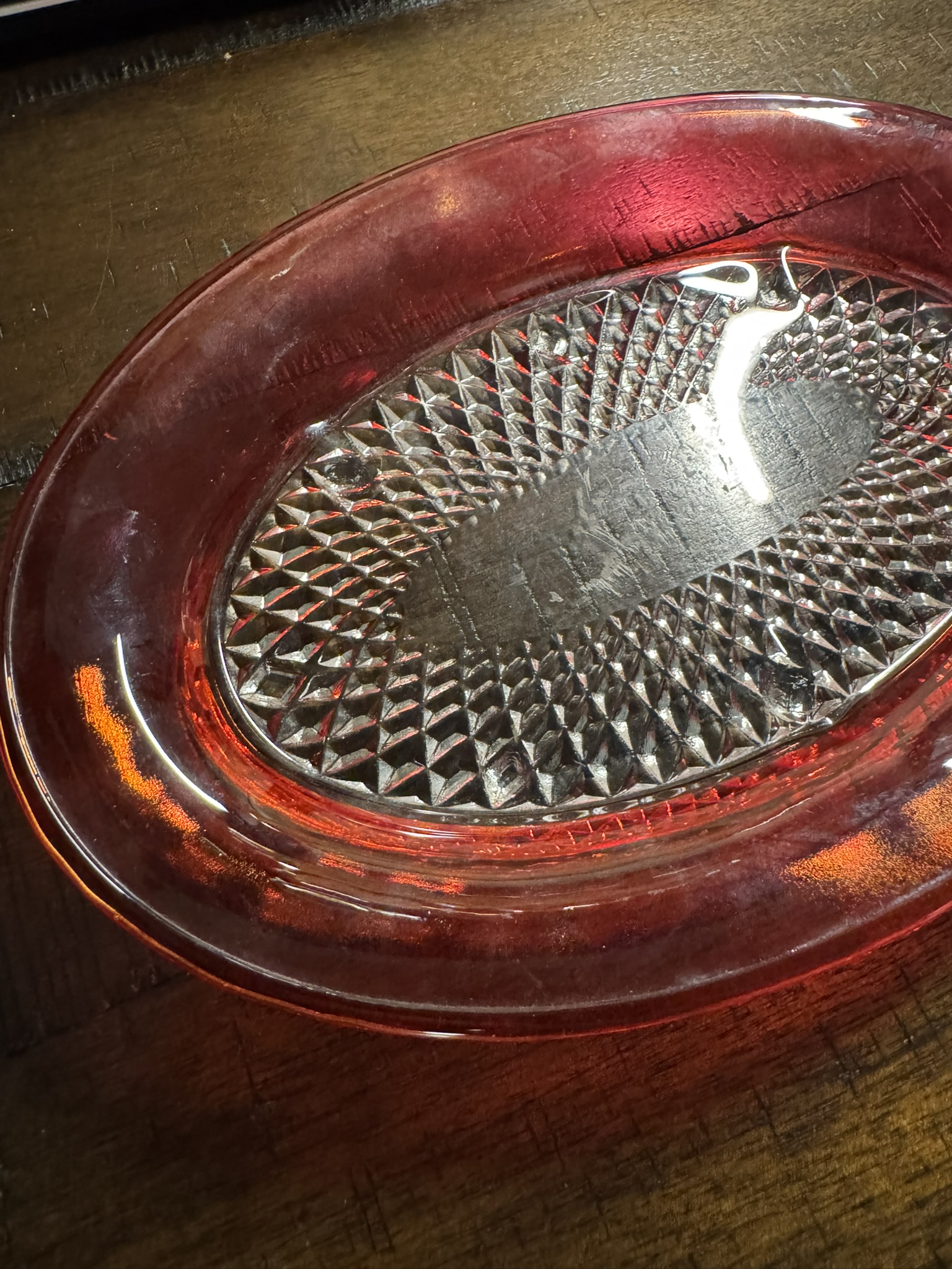 The Ruby Flash Tray is an oval glass tray featuring a transparent red rim and a textured diamond-patterned center. Its glossy finish and intricate design create a warm glow and striking reflections when placed on any surface.