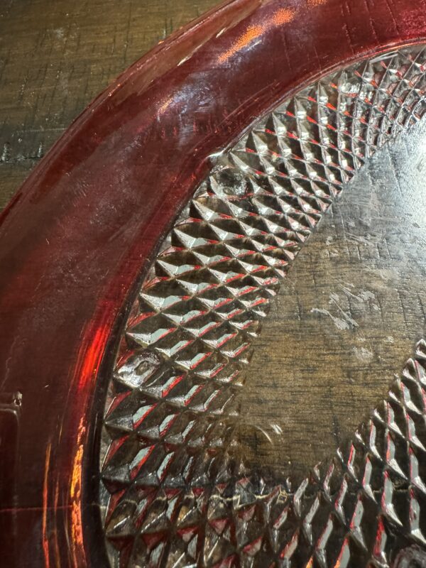 The Ruby Flash Tray, with its textured diamond pattern and smooth curved rim, sits on a worn wooden surface. Its transparent design catches the light, casting subtle reflections and shadows below.