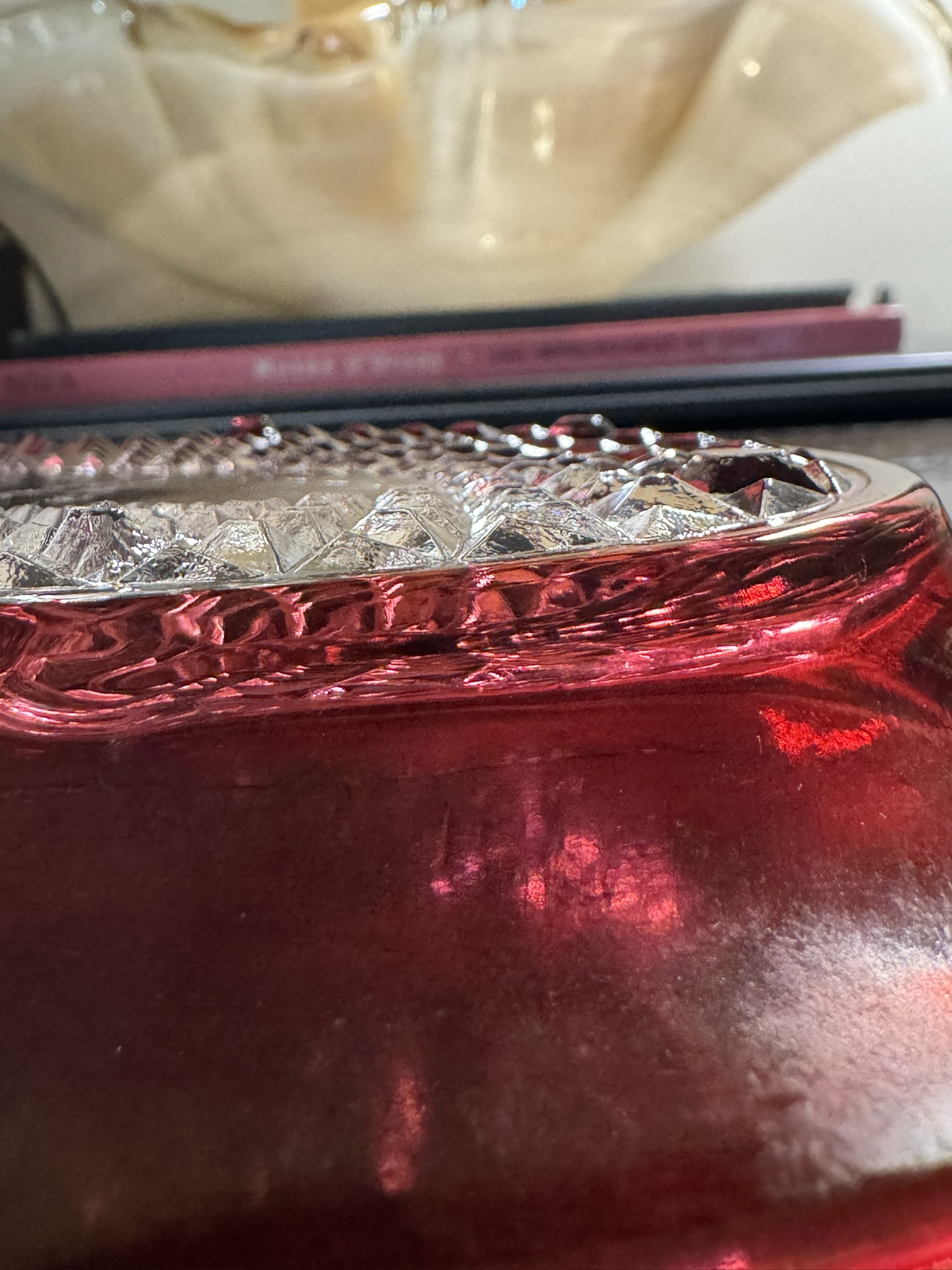 Close-up of the Ruby Flash Tray, featuring a textured, faceted rim that catches the light. A cream decorative object and stacked books appear blurred in the background, accentuating the tray's vivid patterns and brilliant shine.