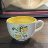 A Stangl Tulip Cup with a yellow interior and painted yellow flowers with green stems sits on a wooden table, while blurred books and a translucent decorative item appear in the background.