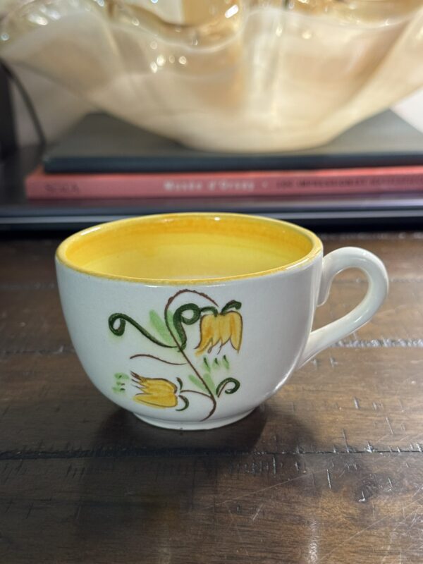 A Stangl Tulip Cup with a yellow interior and painted yellow flowers with green stems sits on a wooden table, while blurred books and a translucent decorative item appear in the background.