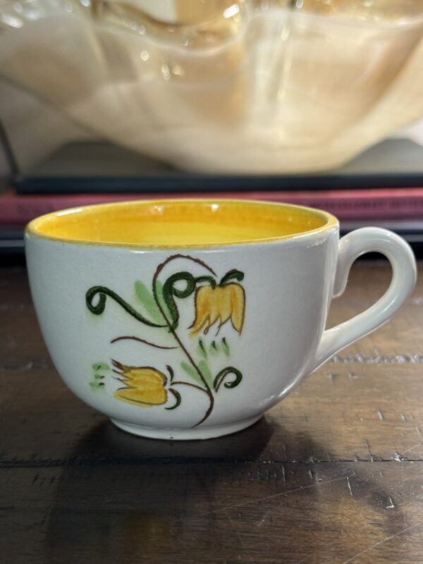 The Stangl Tulip Cup, a ceramic cup featuring a yellow interior and hand-painted tulip design with two yellow flowers and green leaves, sits on wood with books and sheer, light-colored fabric blurred in the background.