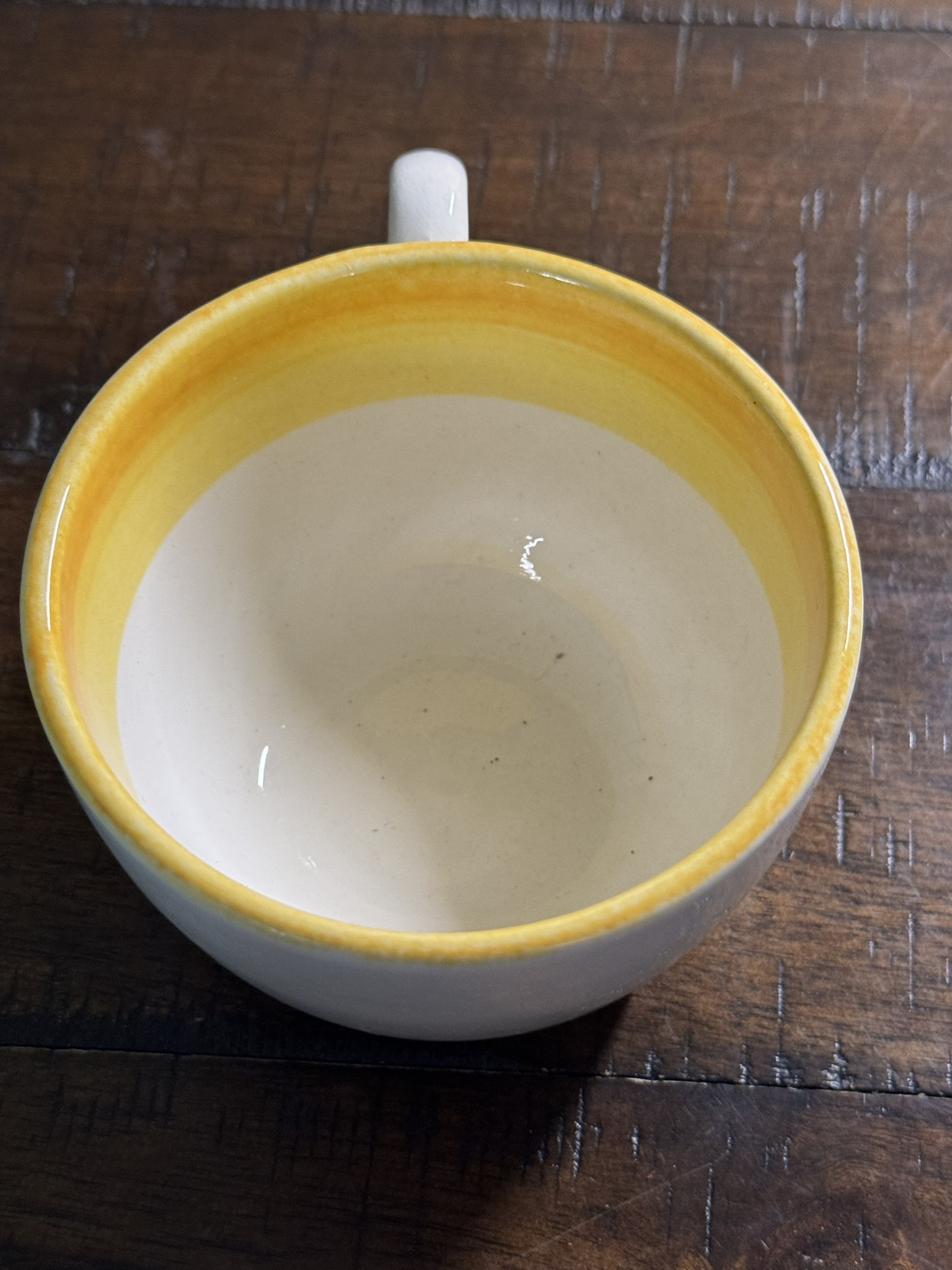 The Stangl Tulip Cup, a white ceramic mug with a yellow rim and gradient yellow band inside, rests on a dark wooden table. Its handle appears at the top of the image, highlighting the cup's rustic charm.