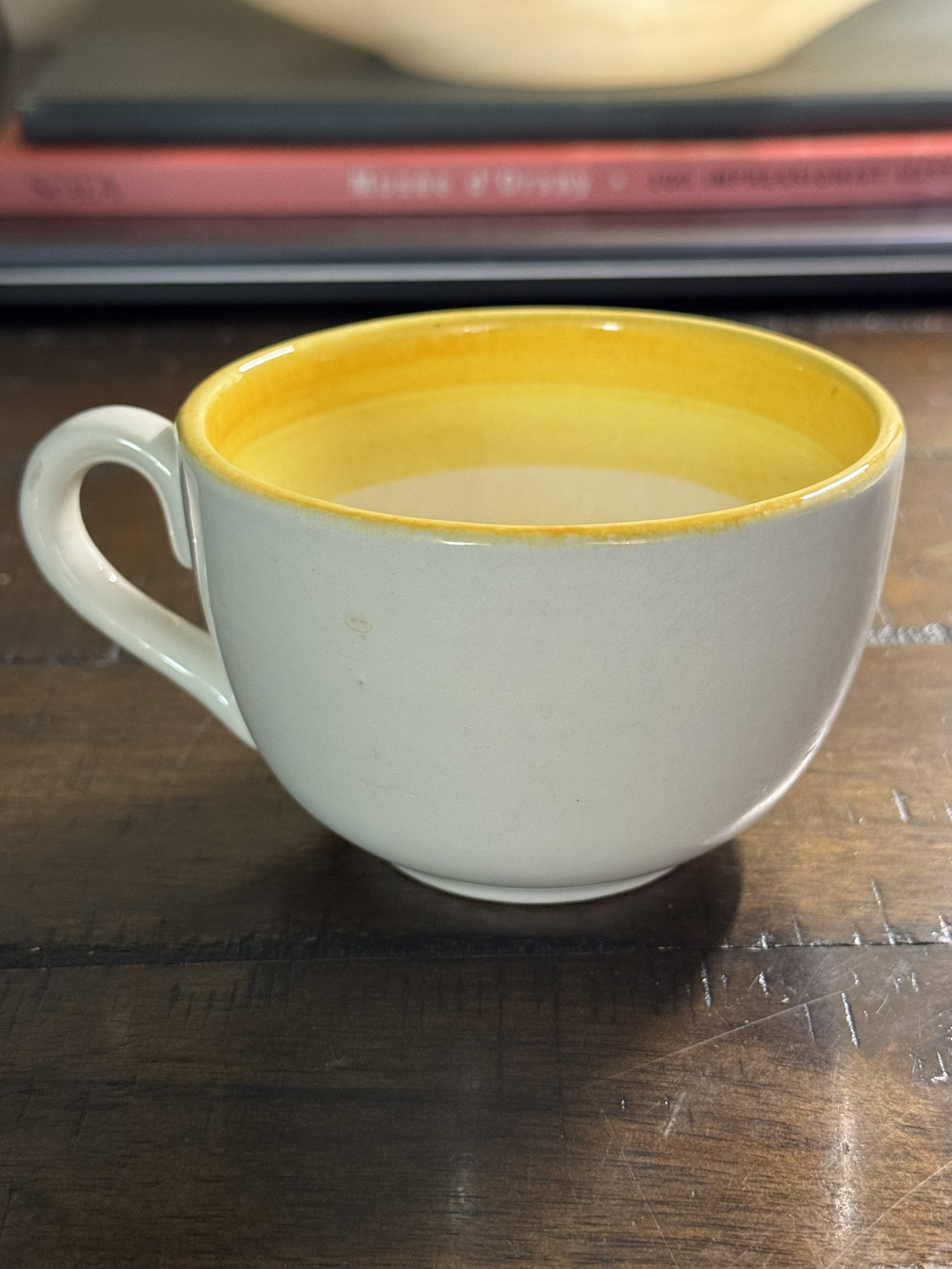 A Stangl Tulip Cup with a yellow rim and small handle rests empty on a dark wooden surface, while stacked red and gray books appear blurred in the warmly lit background.
