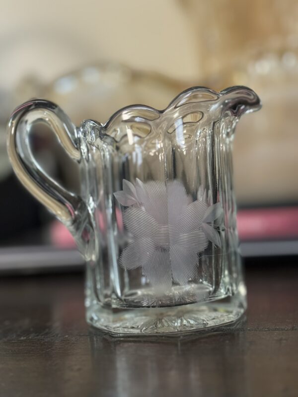 A clear glass creamer pitcher with a decorative, scalloped rim and an etched floral design on the side, sits on a dark surface. The background is softly blurred with warm beige and pink tones.
