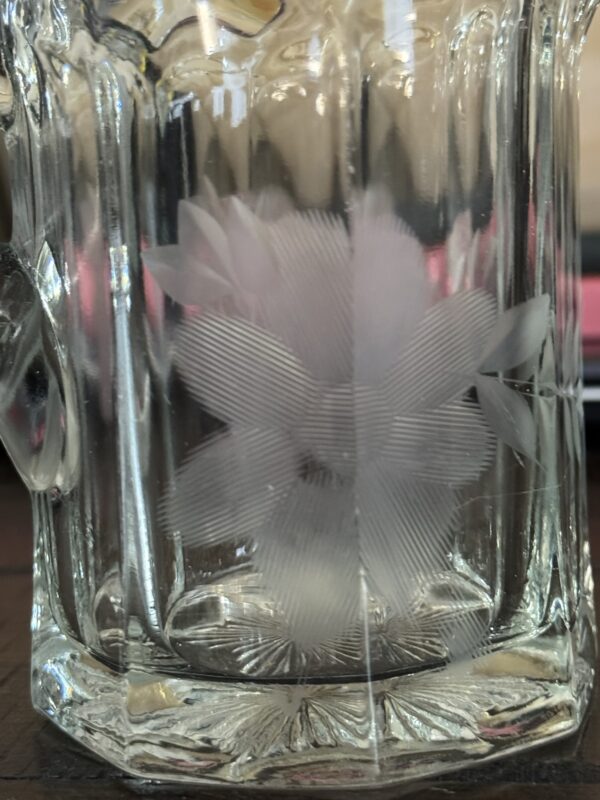 A close-up of a clear glass mug with an etched, frosted flower design on its surface. The flower has delicate petals with fine lines, and the background is softly blurred, showing hints of pink and dark colors.