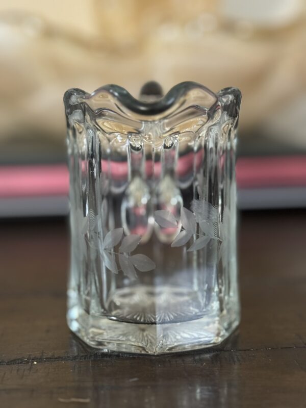 A clear, vintage glass tumbler with ornate, wavy edges, and vertical ridges. The front features an etched design of leaves. The glass sits on a wooden surface with a blurred pink and beige background.