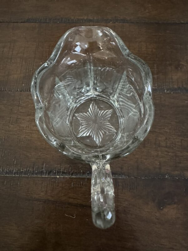 A clear glass cup with a single handle, viewed from above, sits on a wooden surface. The cup has scalloped edges and a starburst pattern embossed on its base. The wood beneath shows visible grain and engraved lines.