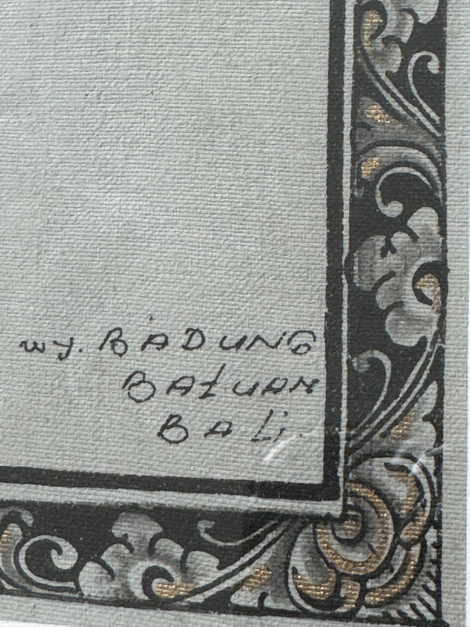 A close-up of a canvas corner shows a decorative black and gold border with floral patterns. Inside the border, handwritten text reads: WJ. RADUNG BATUAN BALI. The background is a light, textured surface.