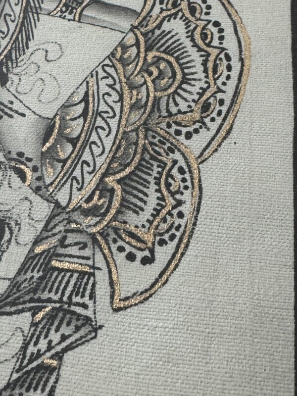 A close-up of an ornate fabric or textile features intricate black and gold patterns with swirling and scalloped details. The textured surface highlights the shimmering gold accents and fine black lines against a light gray background.