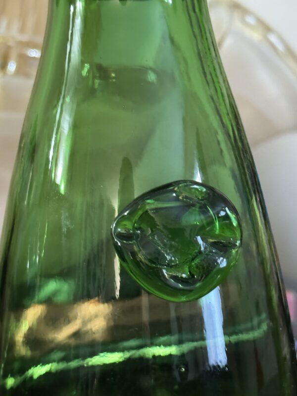 Close-up of the Tuscan Hand-Blown Green Glass Bottle with Seal, highlighting a large round bubble in the smooth green glass. Light reflections and streaks are visible, with a blurred beige background seen through the bottle.