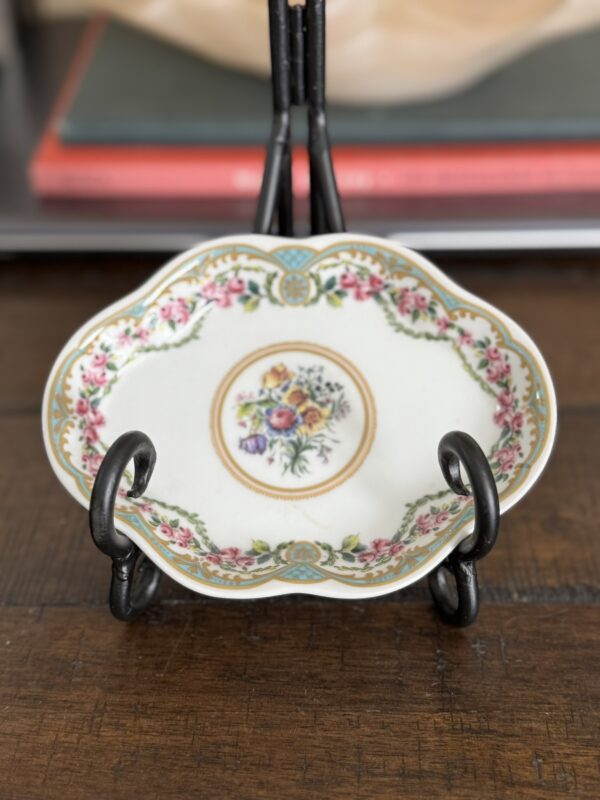 The Sadek Sevres Dish, featuring ornate scalloped edges and a floral center, rests on a decorative black metal stand atop wood. In the background, books with red and gray covers appear blurred.