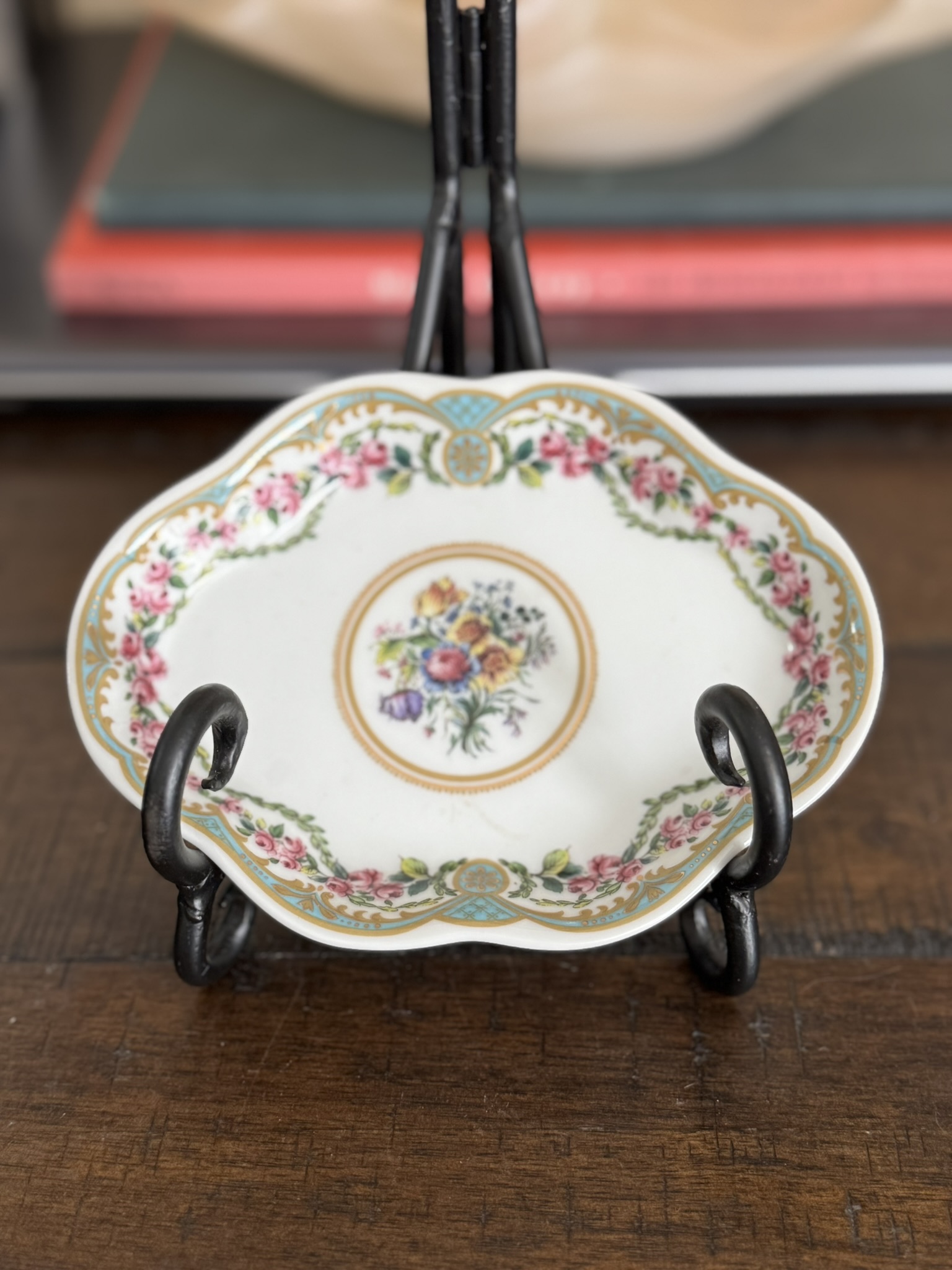 The Sadek Sevres Dish, featuring ornate scalloped edges and a floral center, rests on a decorative black metal stand atop wood. In the background, books with red and gray covers appear blurred.
