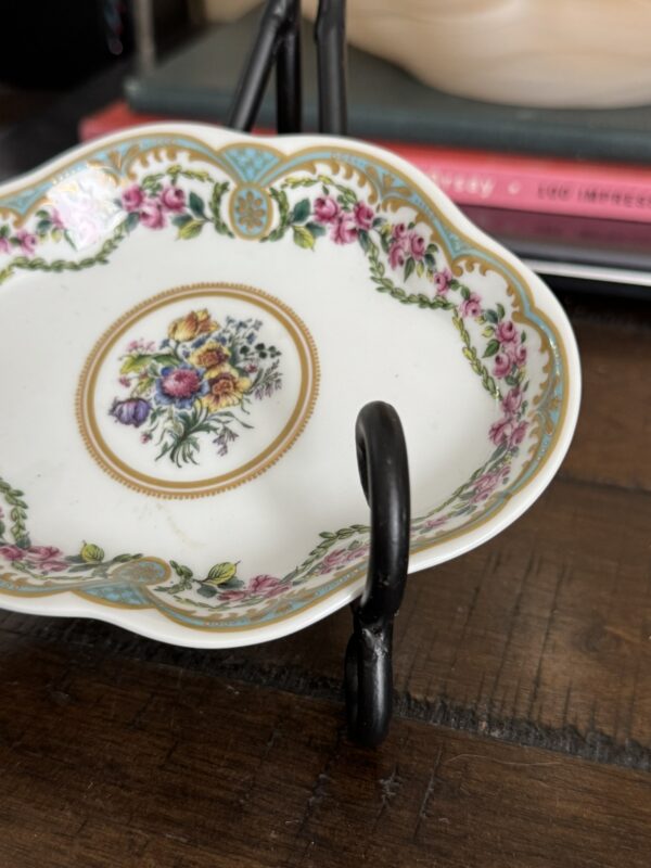 A Sadek Sevres Dish featuring scalloped edges, floral patterns, and gold accents rests on a black metal stand atop a wooden surface, with stacks of colorful books out of focus in the background.