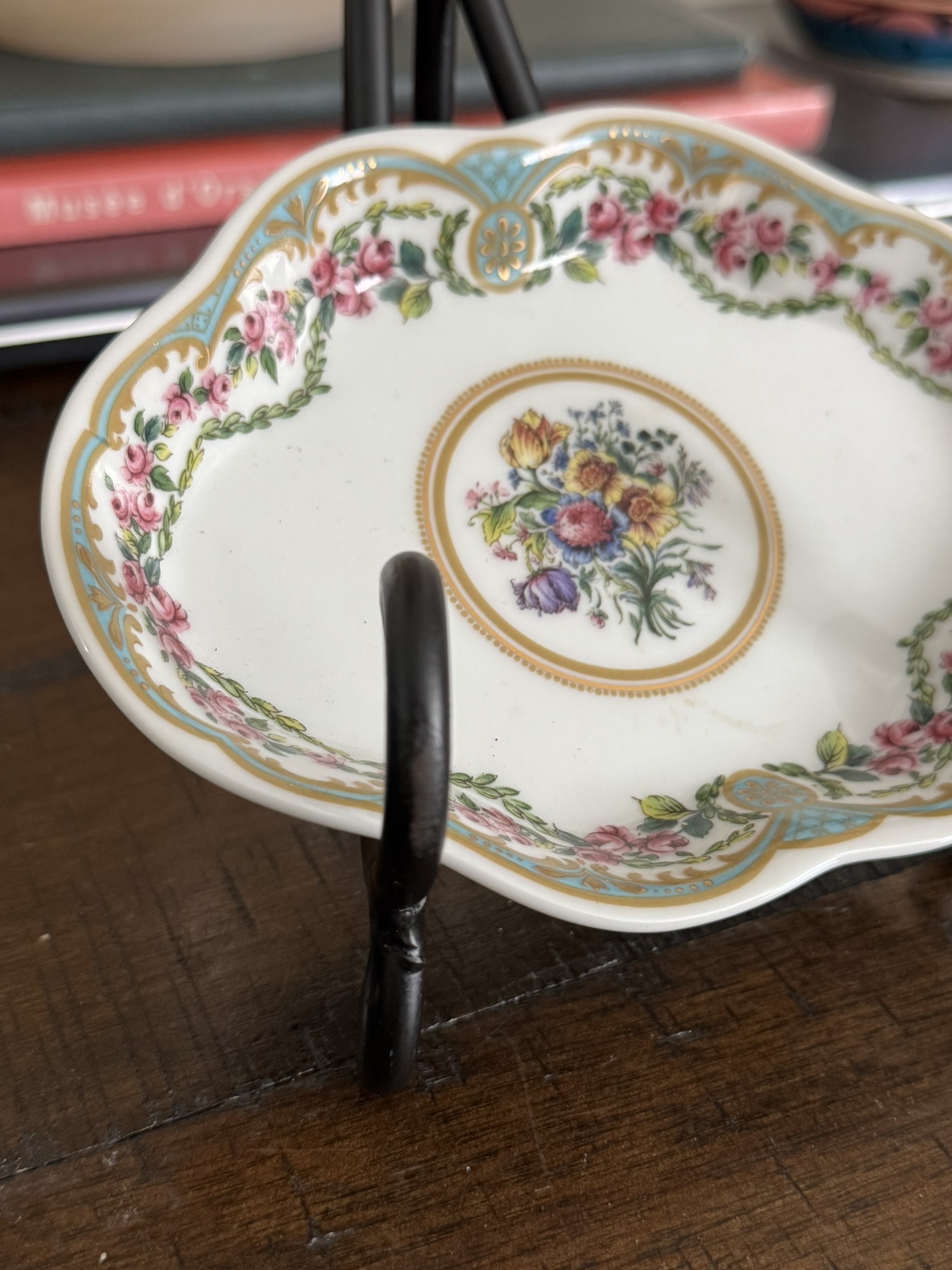 The Sadek Sevres Dish features a scalloped white porcelain design with ornate gold trim and intricate pink, yellow, and purple floral patterns, all beautifully displayed on a black metal stand.