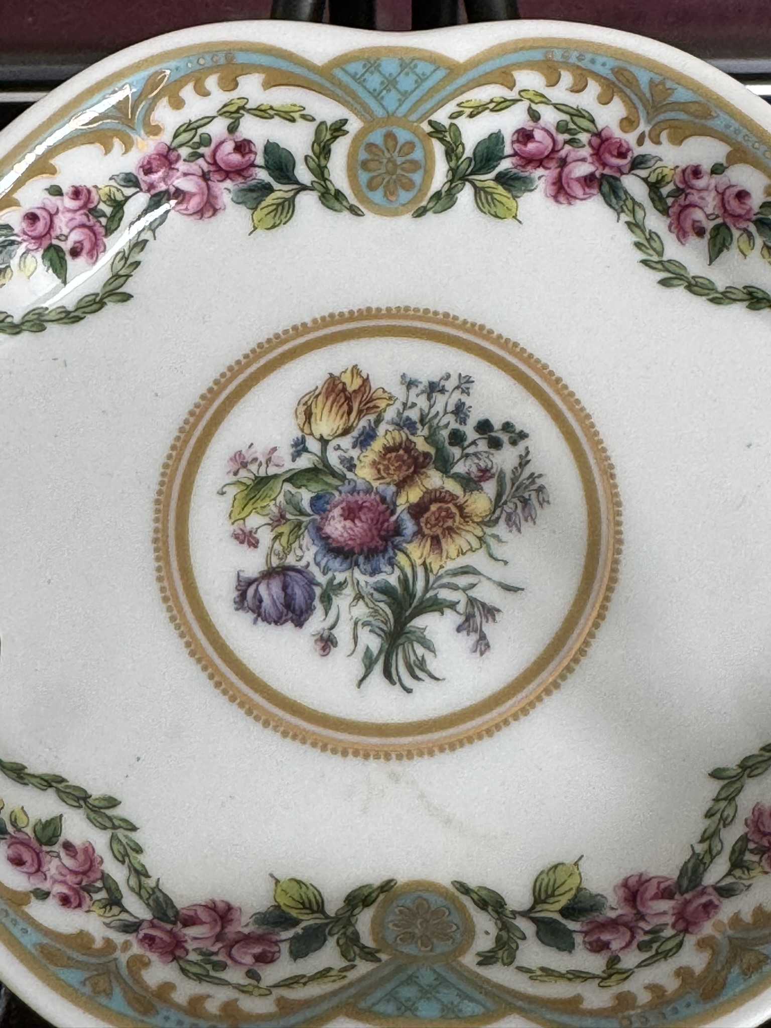 The Sadek Sevres Dish is a decorative porcelain piece featuring a central bouquet of colorful flowers encircled by gold, with a border of pink rose garlands, green leaves, and blue and gold Sevres-style geometric patterns.
