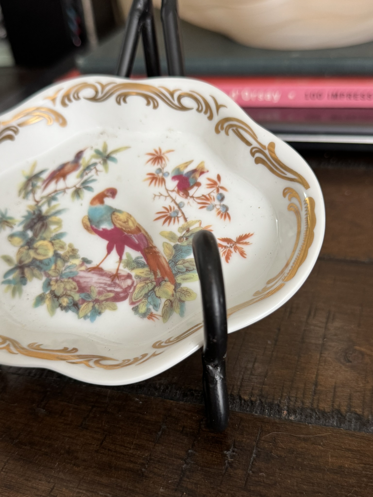 A small white Sadek Bird Dish with gold trim and painted birds and flowers rests on a black metal stand on a wooden surface, with red and white books partially visible in the background.