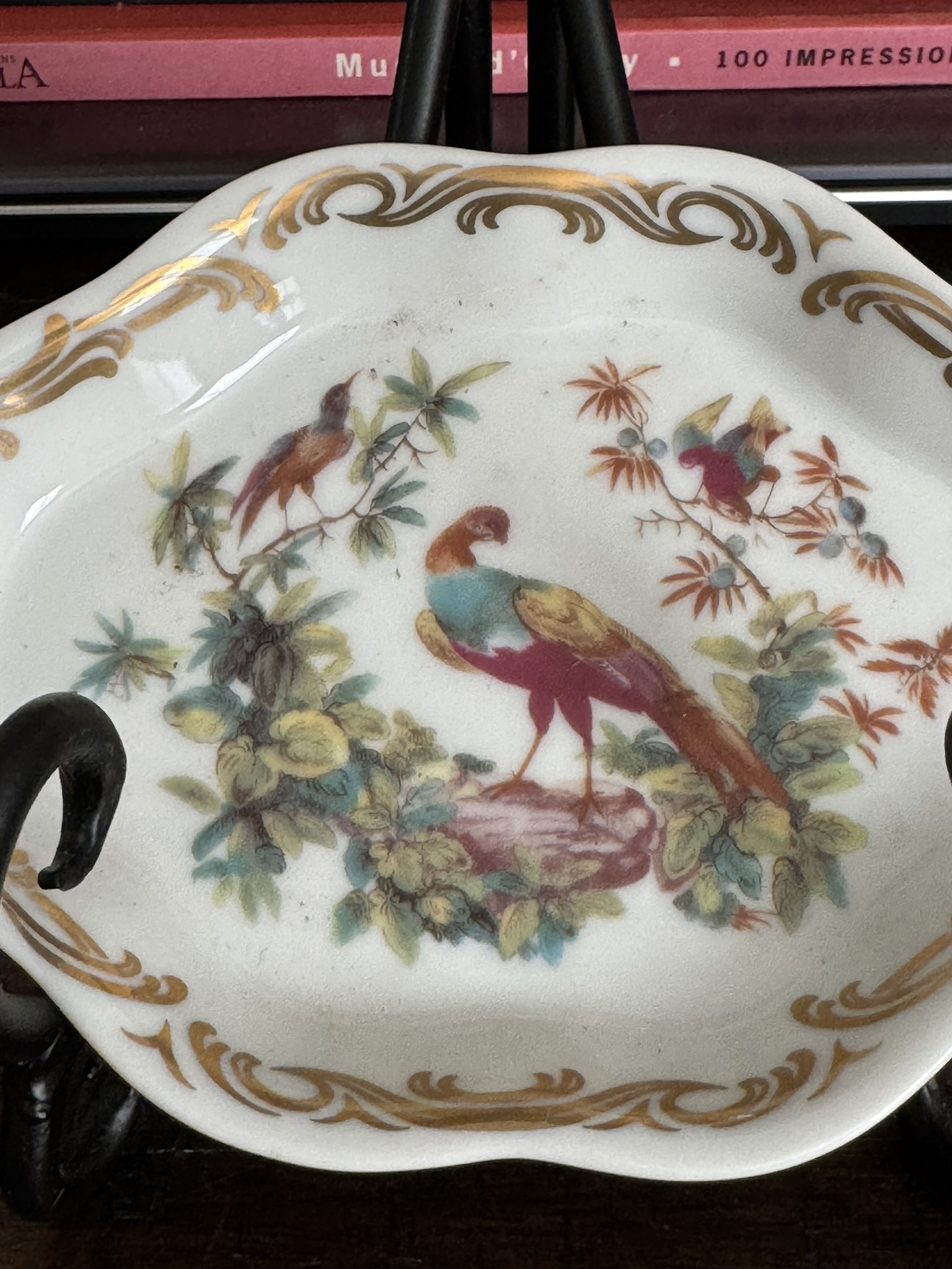 The Sadek Bird Dish is a decorative plate showcasing three colorful birds among green foliage with gold scroll accents on the scalloped rim, displayed on a black metal stand with pink books blurred in the background.