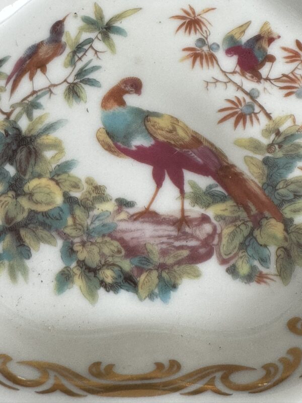 The Sadek Bird Dish features a porcelain design with three birds on leafy branches, one standing on a rock. Ornate gold scrolls accent the border of this colorful and detailed decorative dish.