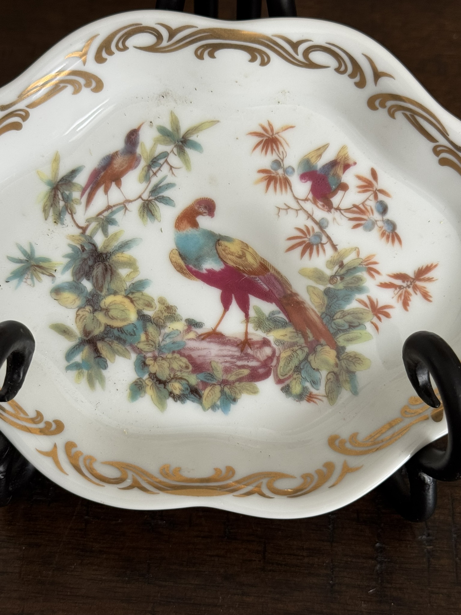 The Sadek Bird Dish is a white porcelain plate with scalloped edges and gold trim, decorated with three colorful birds on leafy branches and flowers. It’s displayed on a black stand on a wooden surface.
