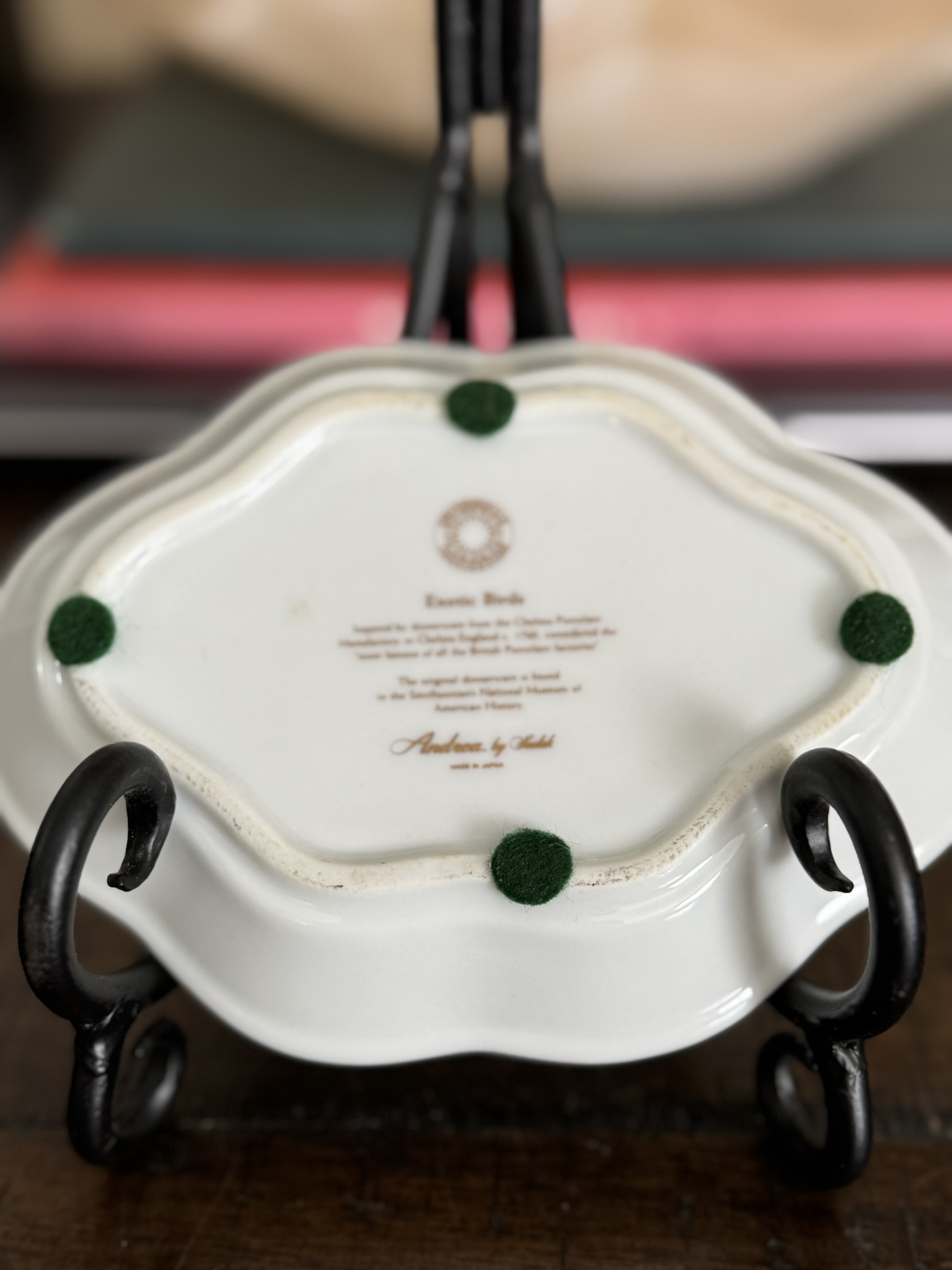 A white Sadek Bird Dish with scalloped edges rests upside down on a black metal stand. Its base shows the maker’s mark, text, and three green felt pads. The background features a blurred brown wooden surface and indistinct objects.