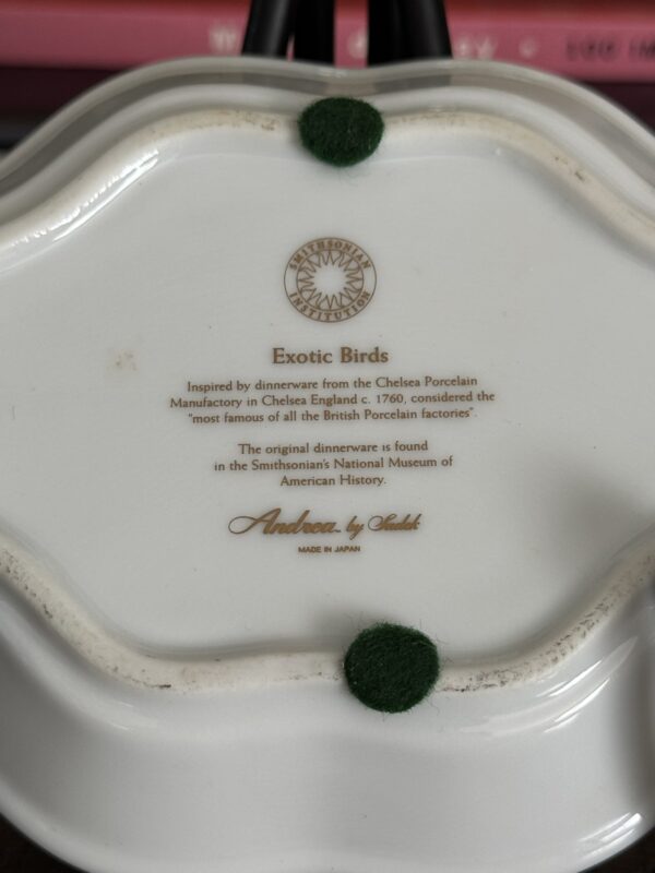 The bottom of the Sadek Bird Dish reads: Sadek Bird Dish, Exotic Birds. Modeled after Chelsea Porcelain Manufactory dinnerware (England, c. 1760), it features a Smithsonian logo, green felt pads, and a pink book blurred in the background.