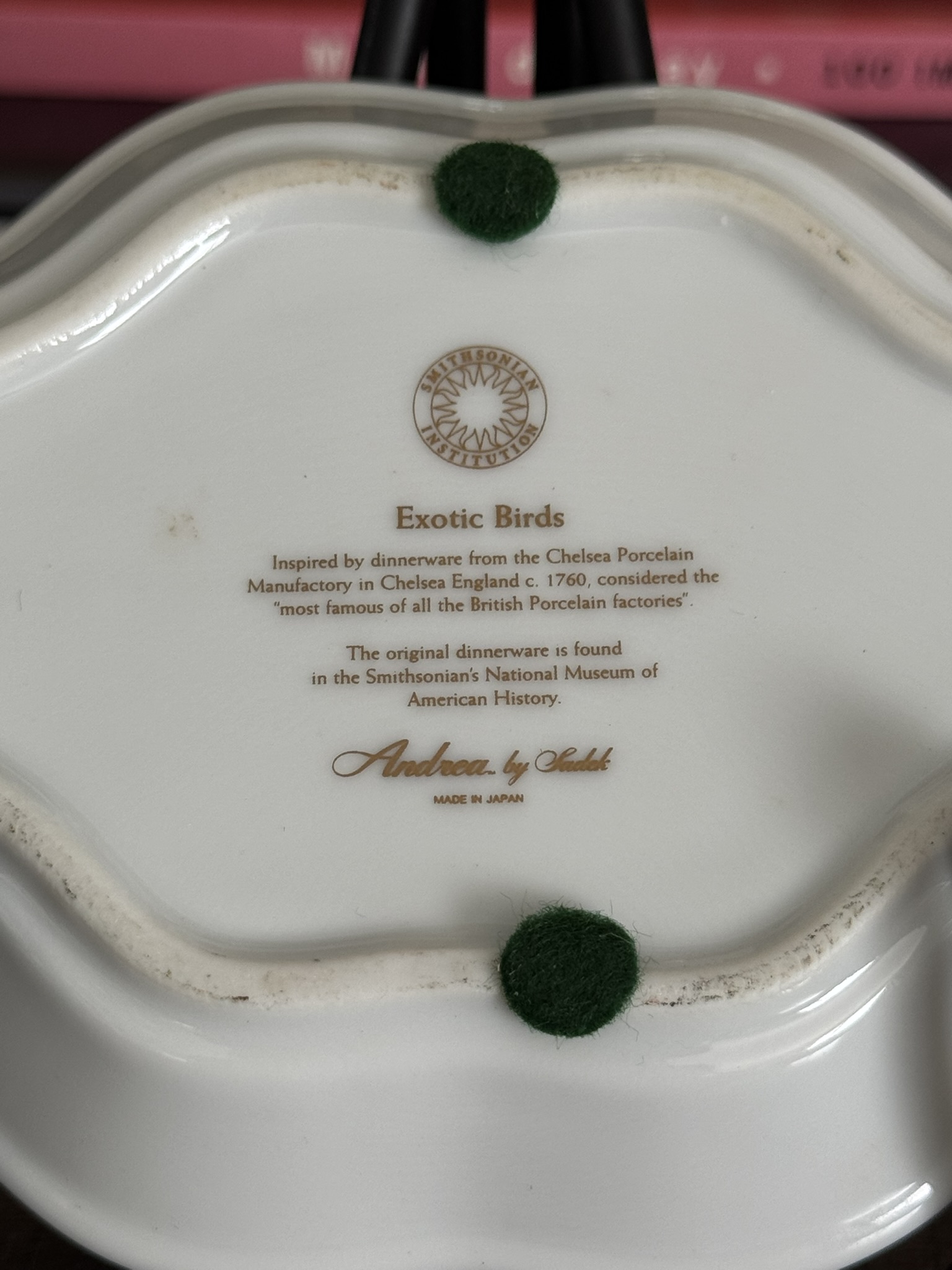 The bottom of the Sadek Bird Dish reads: Sadek Bird Dish, Exotic Birds. Modeled after Chelsea Porcelain Manufactory dinnerware (England, c. 1760), it features a Smithsonian logo, green felt pads, and a pink book blurred in the background.