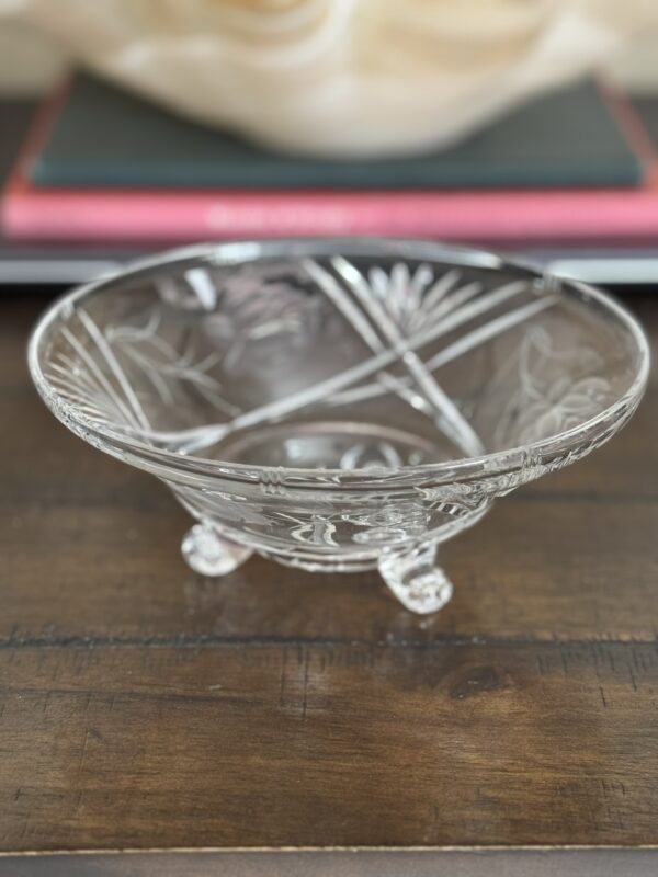 The Vintage Cut Crystal Bowl with Fiddlehead Feet features intricate floral and geometric etching, resting on three ornate feet atop a dark wooden surface, with blurred stacked books and a beige object in the background.