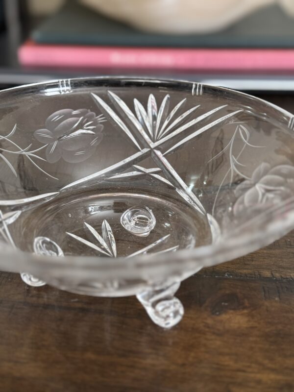 The Vintage Cut Crystal Bowl with Fiddlehead Feet features floral and leaf etchings, intricate engraved designs, and rests on a wooden surface. Books with red and black spines are partially visible in the blurred background.