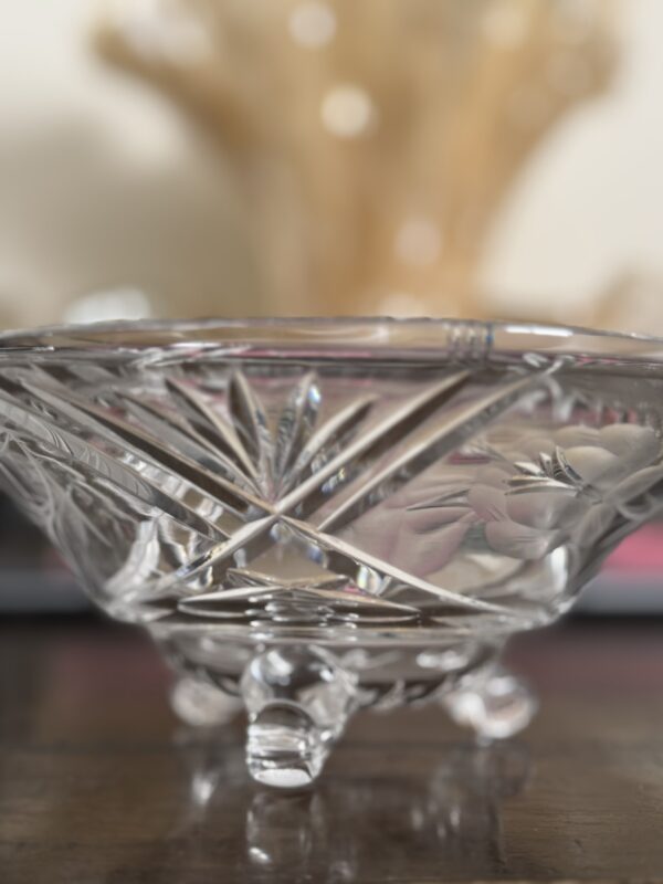 The Vintage Cut Crystal Bowl with Fiddlehead Feet features a clear, detailed starburst and crisscross pattern. It stands on three ornate feet atop a wooden surface, set against a soft warm beige and gold background.