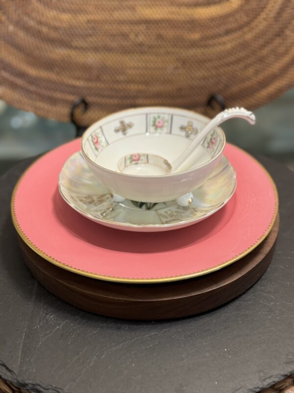 A stack of dishes sits on a dark surface: a pink plate with gold rim, smaller brown plate, white saucer, and on top, the Sadek Bird Dish teacup and matching bowl with ladle. A woven placemat stands behind.