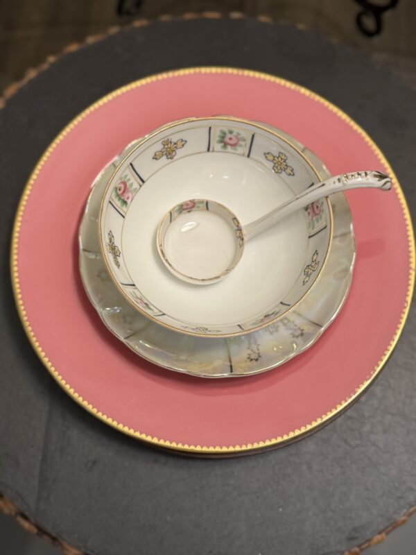 A porcelain bowl with a matching spoon rests inside a scalloped saucer, all set atop a large pink plate with a gold beaded rim. The bowl features floral and geometric designs in soft pastel colors. The stack sits on a dark, round tabletop.