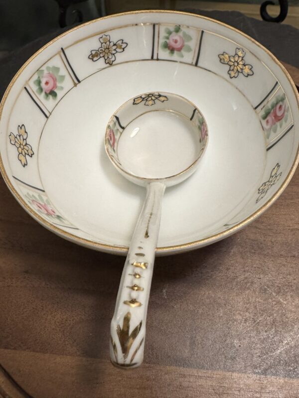 A white porcelain bowl and ladle with gold trim and floral patterns featuring pink roses and yellow flowers. The set sits on a wooden surface, with the ladle resting inside the bowl and its handle extending out toward the viewer.