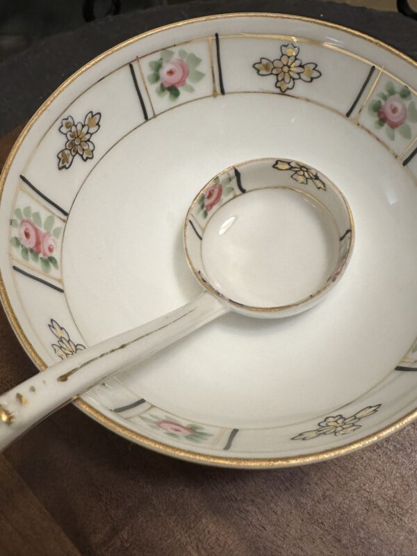 A close-up of a decorative white porcelain bowl and matching ladle, both with gold trim. The bowl features a pattern of pink roses, green leaves, and yellow flowers in panels around the rim. The ladle rests inside the bowl, displaying similar designs.